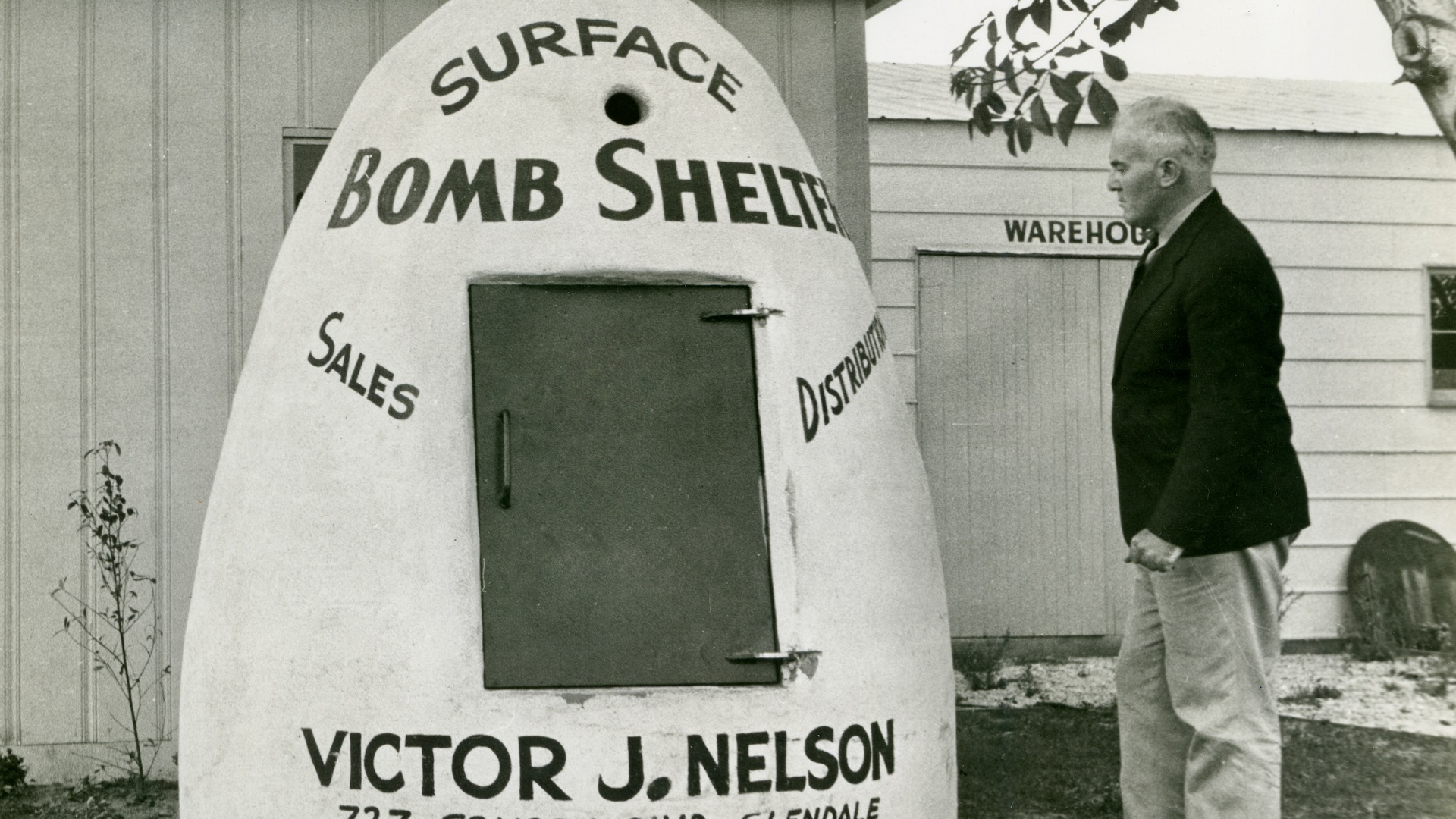 A black-and-white photo shows a man standing next to a surface (above ground) bomb shelter that is advertising Victor J. Nelson's sales and distribution business, circa 1951. (Jim Heimann Collection/Getty Images)