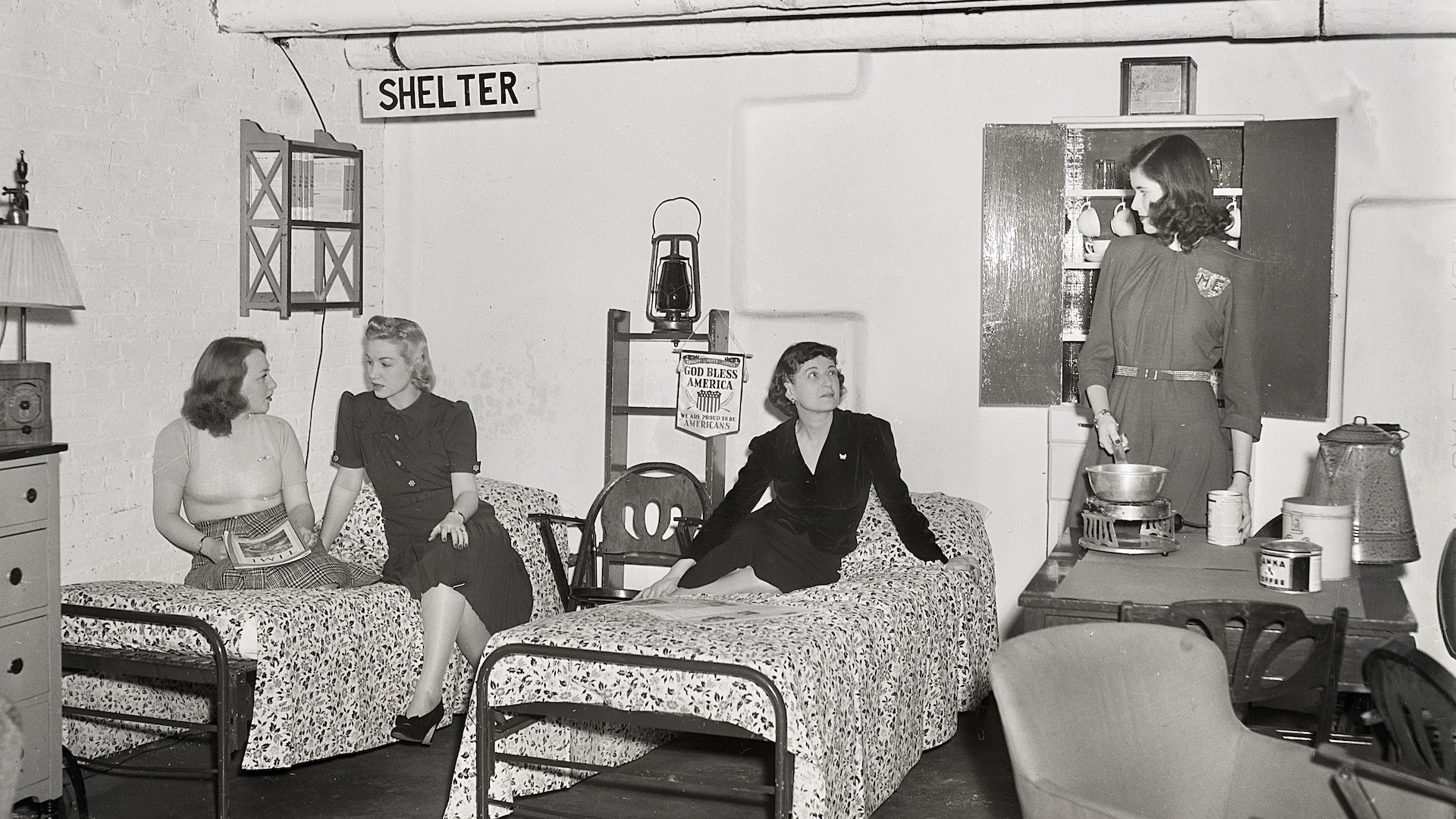 3/12/1941- New York, NY: Hotel completes air raid shelter. A few of the Allerton House's guets showed how comfortable and practical are the conveniences incorpoarted in the new air-raid shelter completed in the sub-basement of the hotel. The shelter is approximately 45 feet underground, and an auxiliary lighting system has been installed in case the hotel's own light plant goes out of order. Here, four women are shown cooking and reclining on the bomb shelter's beds. A bannerhung on the wall next to a hurricane lamp says "God Bless America."