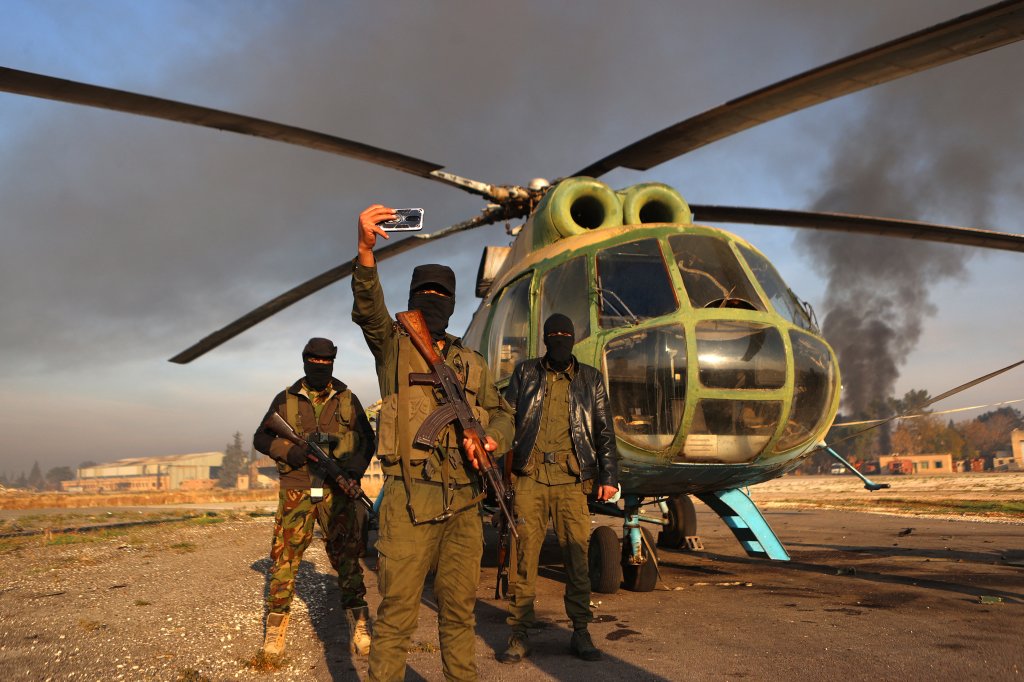 TOPSHOT - Anti-regime fighters pose for a picture with an army helicopter on the tarmac at the Nayrab military airport in the northern Syrian city of Aleppo on December 2, 2024. Islamist-led rebels on November 30, seized most of Aleppo, along with its airport and dozens of nearby towns, the war monitor Syrian Observatory for Human Rights said. (Photo by AAREF WATAD / AFP) (Photo by AAREF WATAD/AFP via Getty Images)