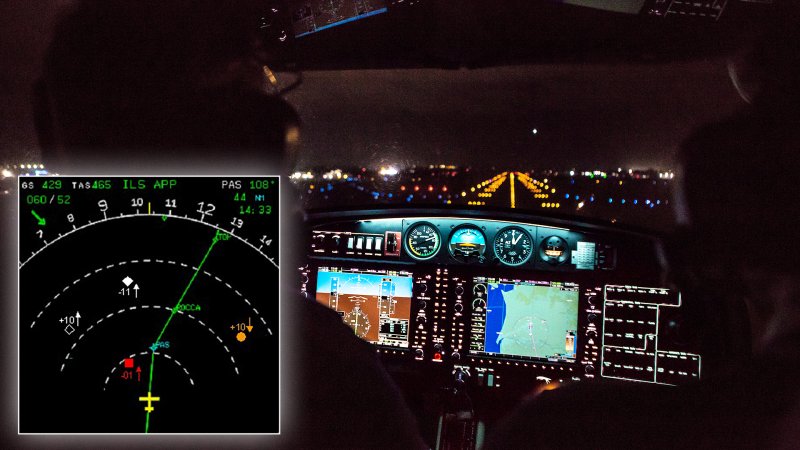 A caucasian uniformed pilot in the cockpit of a small, modern private single engine propeller aircraft landing at night with runway lights visible.